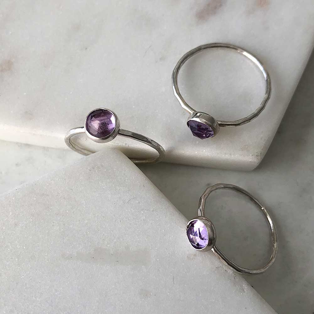 Three silver rings with hammered bands and bezel-set amethysts displayed on marble. 