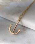 A gold-filled, loosely anchor-shaped pendant on a fine chain, displayed on marble. 