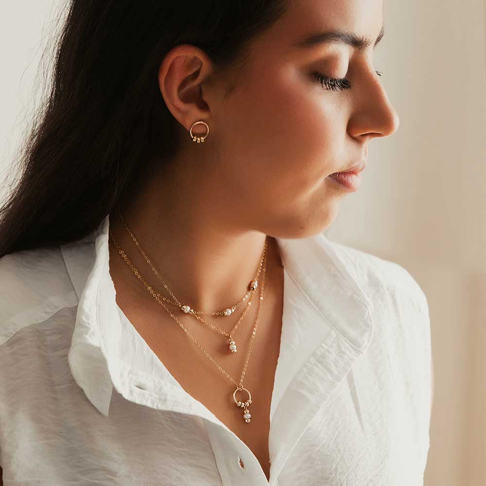 A gold-filled circle stud earring with three polished gold-filled beads, worn by a model. The model also wears a selection of Strut necklaces.