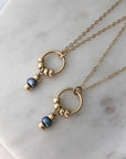 Two circle pendant necklaces, each with four polished gold-filled beads and a hanging attachment with two beads and a dark pearl. Hung from fine gold-filled chain and displayed on marble. 