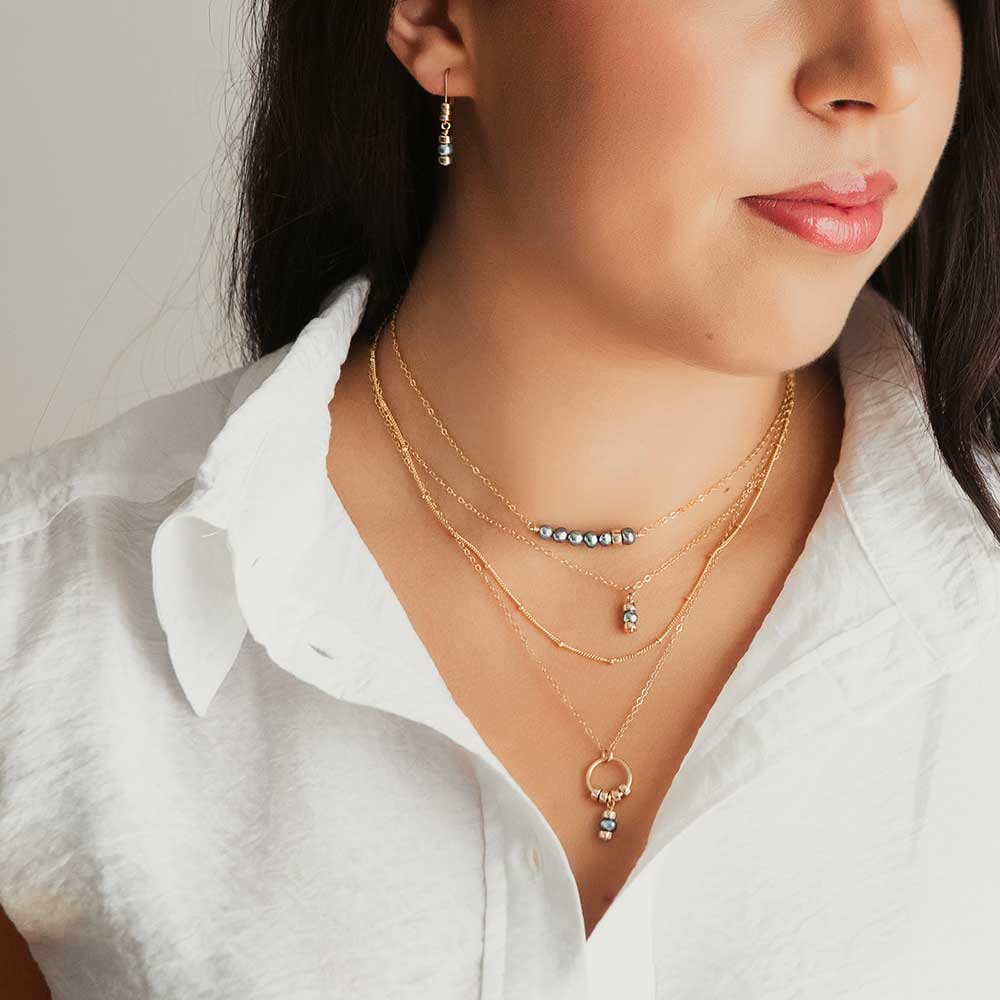 A bar pendant with irregularly shaped dark pearls and a single polished gold-filled bead, attached on either side to fine gold-filled chain. Worn with either necklaces by a model. Hangs close to the throat. 