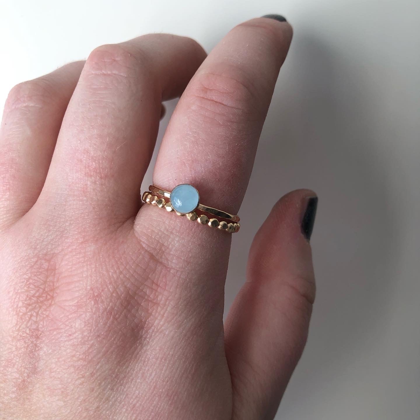 Two rings worn by a model with dark nailpolish. One is a gold-filled beaded band. The other is a hammered gold-filled band set with a pale blue aquamarine in a bezel. 