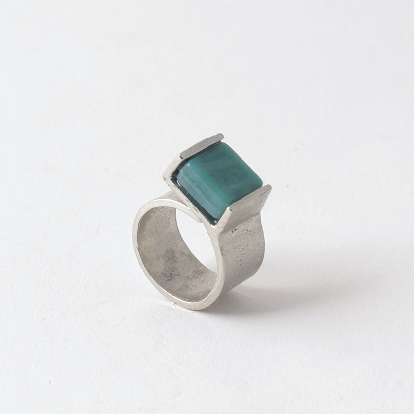 Pictured: Bello ring in &#39;Aquatic&#39; against flat white background. The ring is a wide flat strip of pewter that curls back on itself but is not soldered. At the end of the overlap, a square piece of green-blue opaque glass has been set in a half-bezel.