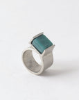 Pictured: Bello ring in 'Aquatic' against flat white background. The ring is a wide flat strip of pewter that curls back on itself but is not soldered. At the end of the overlap, a square piece of green-blue opaque glass has been set in a half-bezel.