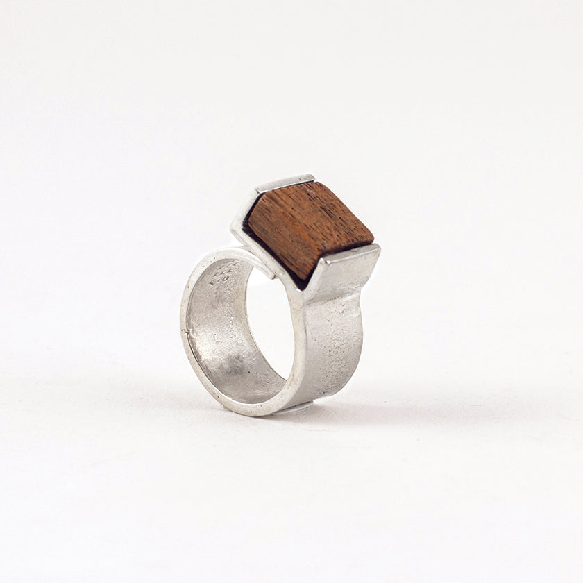 Pictured: Bello ring in 'Wood' against flat white background. The ring is a wide flat strip of pewter that curls back on itself but is not soldered. At the end of the overlap, a square piece of dark, warm-coloured wood has been set in a half-bezel.