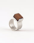 Pictured: Bello ring in 'Wood' against flat white background. The ring is a wide flat strip of pewter that curls back on itself but is not soldered. At the end of the overlap, a square piece of dark, warm-coloured wood has been set in a half-bezel.
