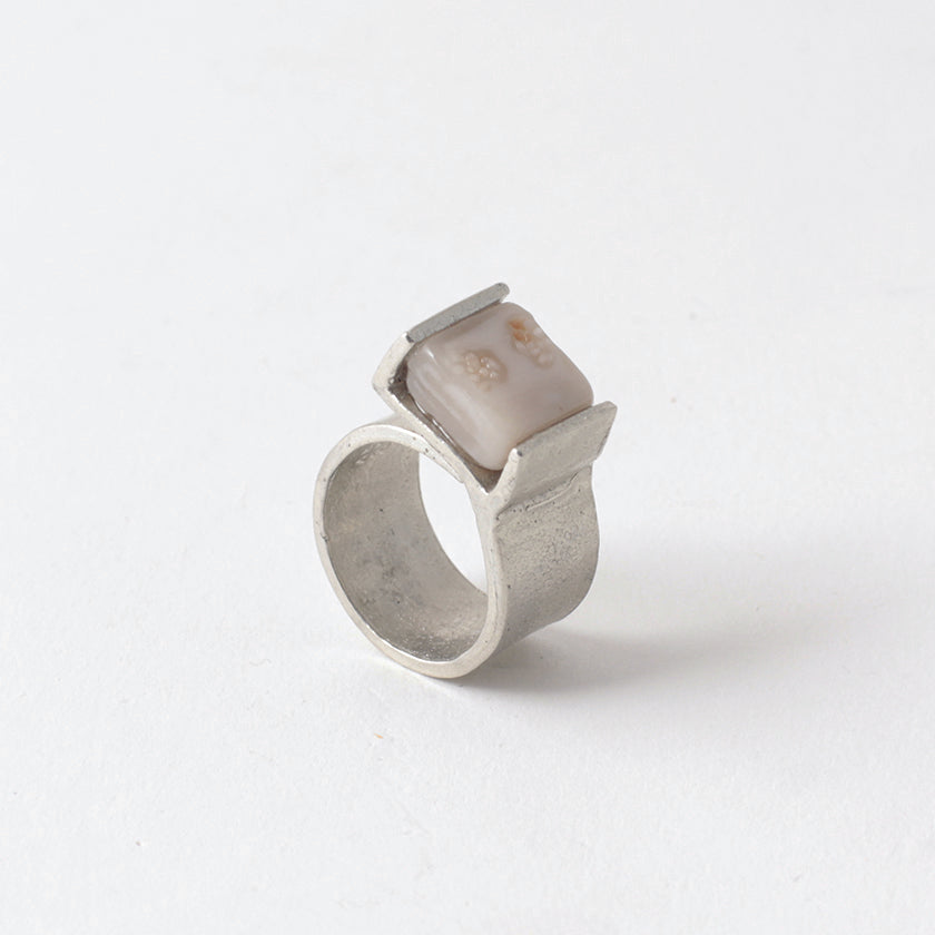 Pictured: Bello ring in 'Sand' against flat white background. The ring is a wide flat strip of pewter that curls back on itself but is not soldered. At the end of the overlap, a square piece of pale, cream-coloured opaque glass with a slight texture has been set in a half-bezel.