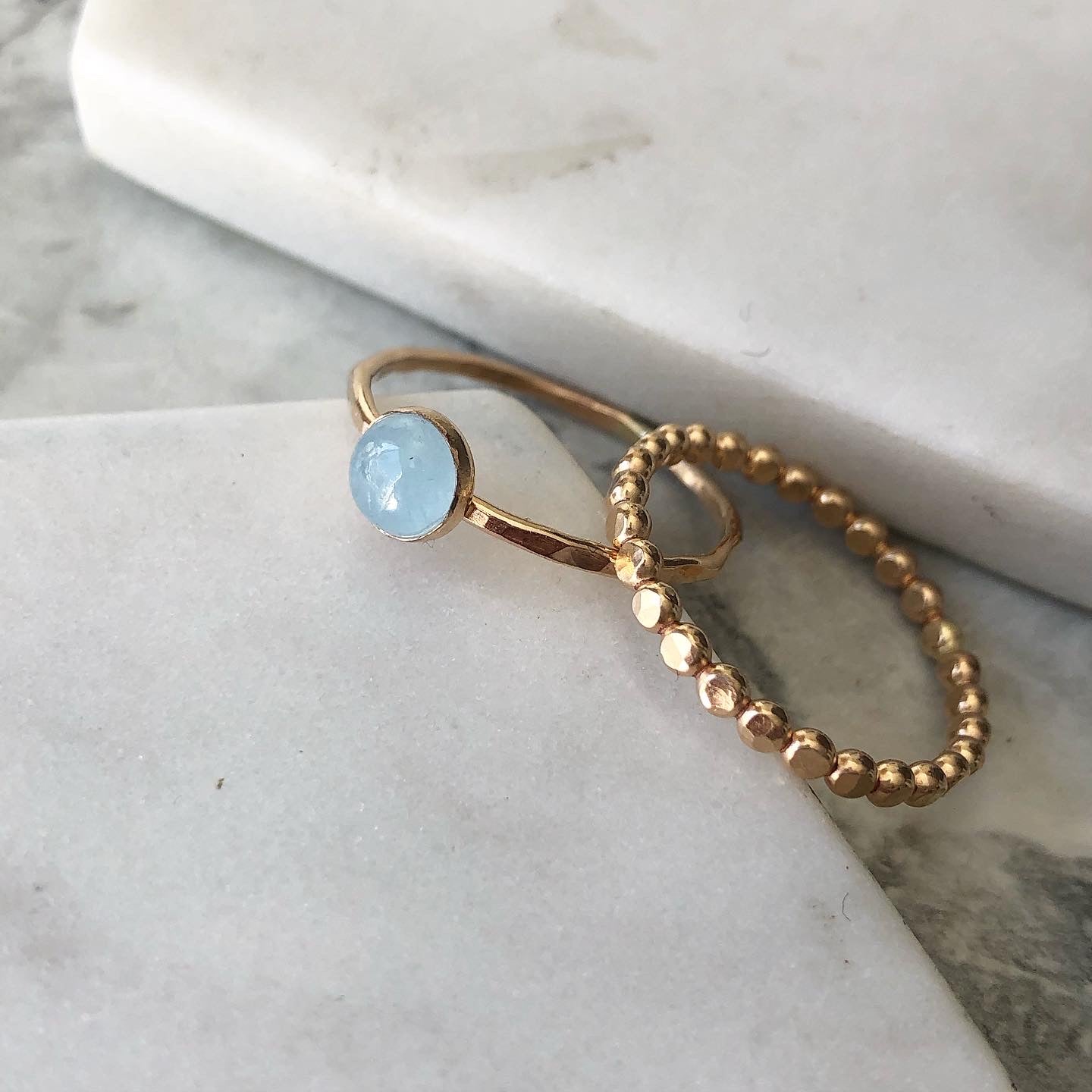 Two rings displayed on marble. One is a beaded gold-filled band. The other is a hammered gold-filled band set with a round pale blue aquamarine in a bezel. 