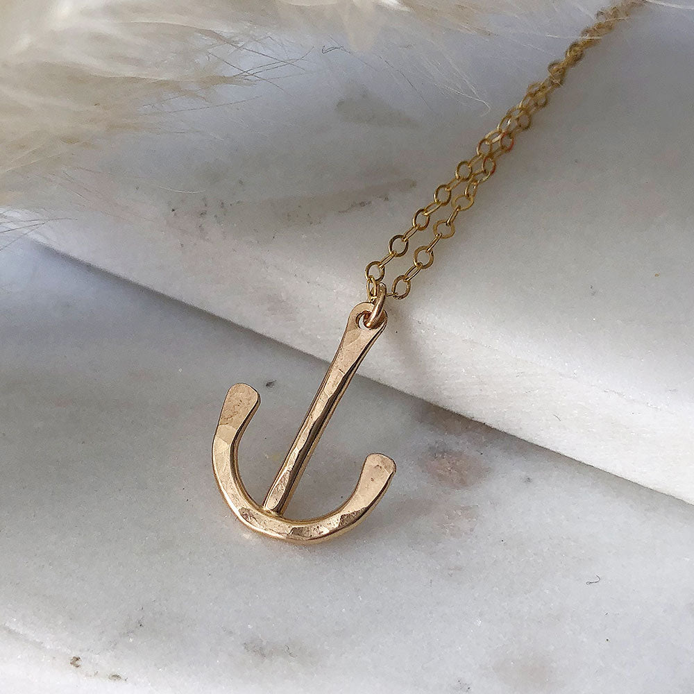 A gold-filled, loosely anchor-shaped pendant on a fine chain, displayed on marble. 
