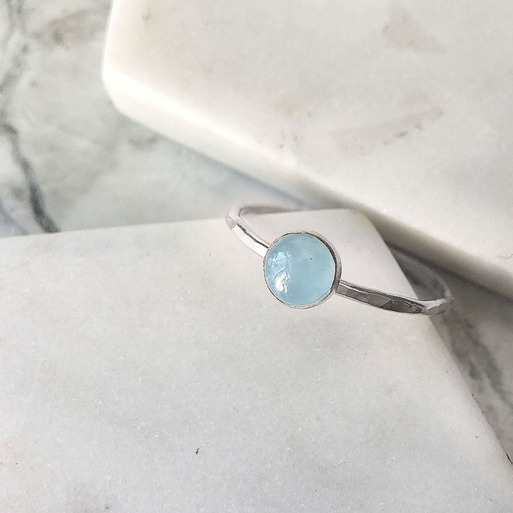 A hammered silver band set with a pale blue aquamarine in a bezel. Displayed on marble.