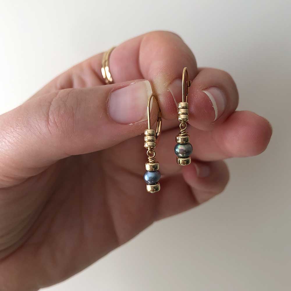 Dark pearl drop earrings on gold-filled leverback hooks embellished with three polished gold-filled beads each. Held by model. 