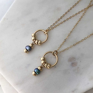 Two circle pendant necklaces, each with four polished gold-filled beads and a hanging attachment with two beads and a dark pearl. Hung from fine gold-filled chain and displayed on marble.  | Magpie Jewellery