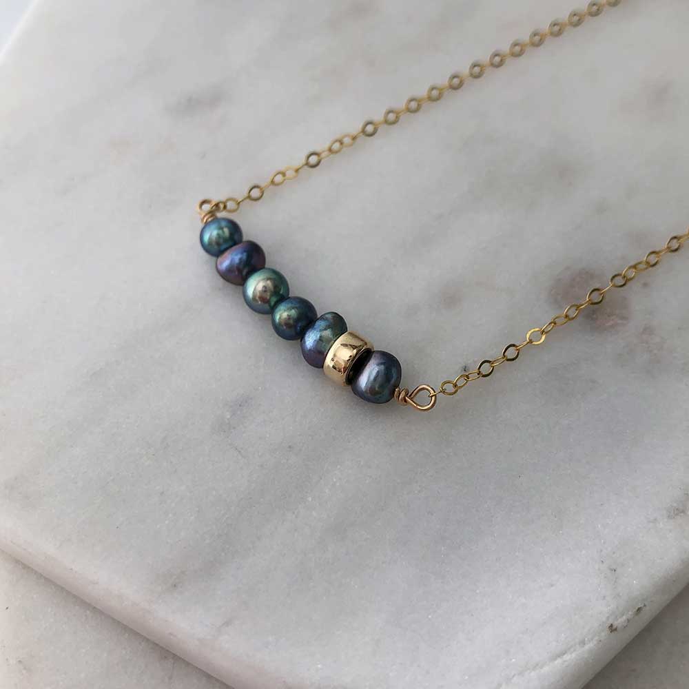 A bar pendant with irregularly shaped dark pearls and a single polished gold-filled bead, attached on either side to fine gold-filled chain. Displayed on marble. 