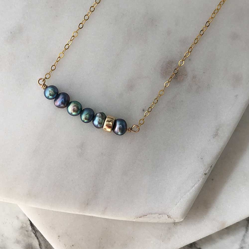 A bar pendant with irregularly shaped dark pearls and a single polished gold-filled bead, attached on either side to fine gold-filled chain. Displayed on marble. 